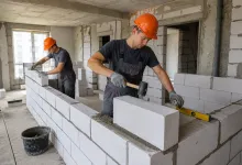 Двоє будівельників у помаранчевих касках укладають блоки з газобетону під час зведення стін у приміщенні